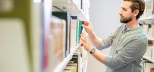 Ein Student steht in der Bibliothek am Bücherregal und sucht ein Buch.