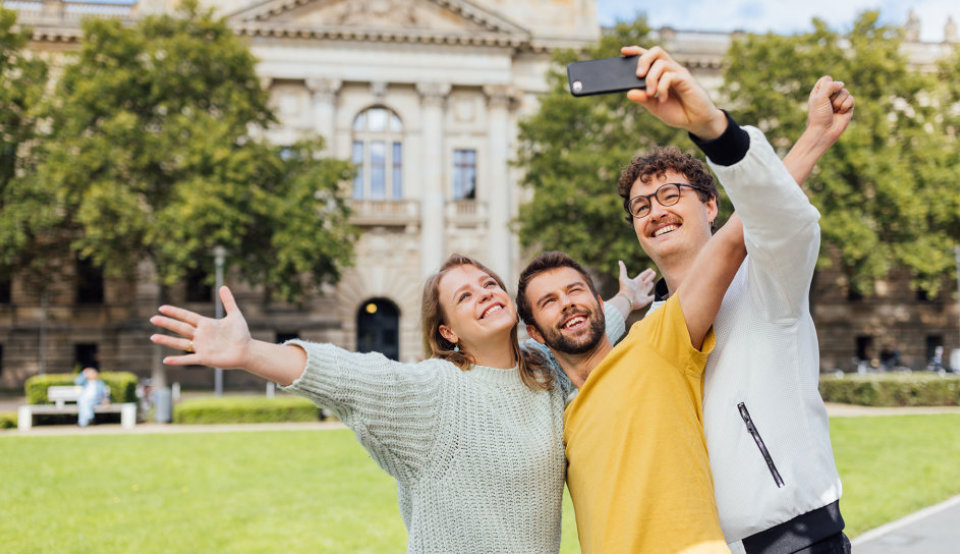 Drei Studierende nehmen ein Selfie mit einem Smartphone auf dem Uni-Gelände auf.