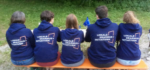 Fünf LEI-Mitglieder sitzen auf einer Bierbank mit dem Rücken zur Kamera und präsentieren das Logo der Lokalen Erasmus+ Initiativen auf der Rückseite ihrer dunkelblauen LEI-Hoodys.