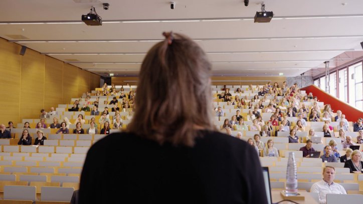 Blick auf den Rücken der Rednerin in den Hörsaal mit Publikum