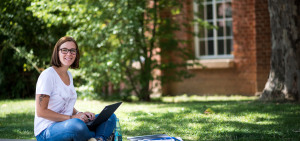Studierende sitzt im Schneidersitz mit Laptop auf den Knien auf Grünfläche eines Uni-Geländes