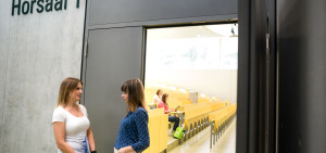 Zwei Studierende stehen vor der offenen Türe eines Hörsaals und unterhalten sich.