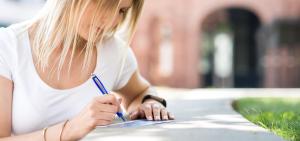 Studentin sitzt im Freien an einer Mauer und notiert sich etwas auf einer Karte
