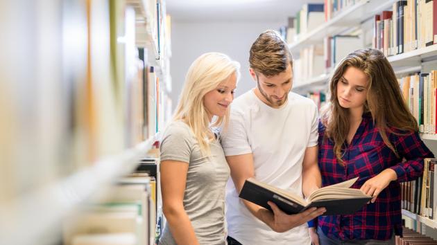 Drei Studierende lesen zwischen Bibliotheksregalen ineinem Buch