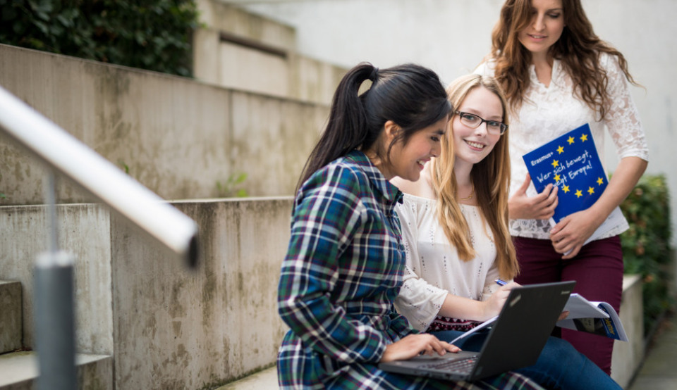Drei Studentinnen unterhalten sich im Freien auf einer Treppe über das Jean Monnet Programm, während eine von ihnen nach den Aufnahmebedingungen auf ihrem Laptop recherchiert.