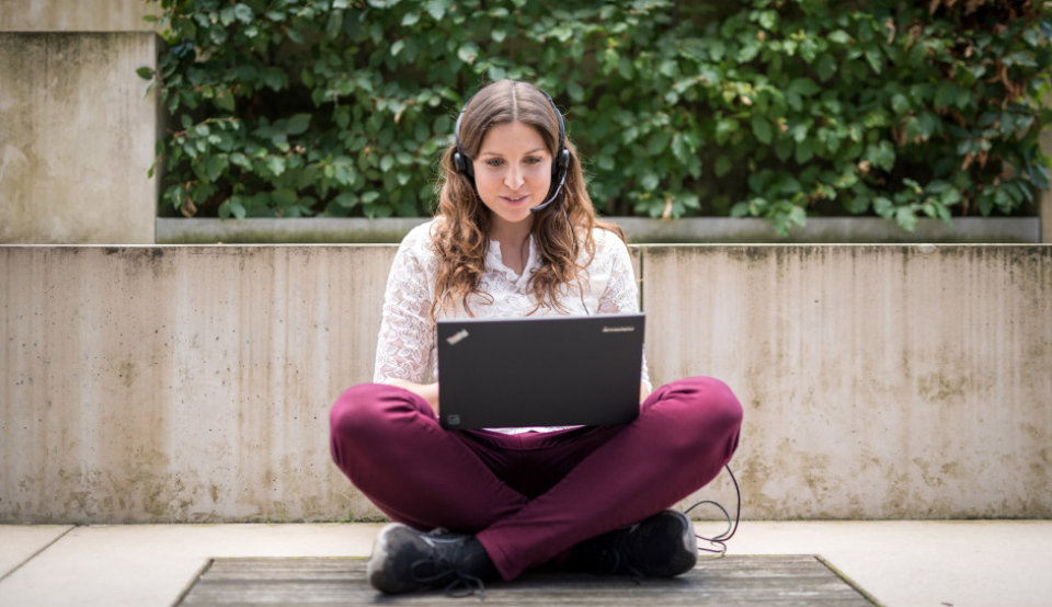 Eine Studentin arbeitet mit Headset im Schneidersitz an ihrem Laptop