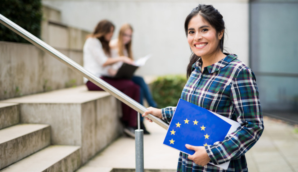 Eine Studentin hält ein Prospekt der NA DAAD mit der europäischen Flagge im Arm. Zwei Studierende informieren sich über die Erasmus+ Partnerländer.