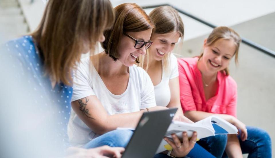 Vier Studierende unterhalten sich im Uni-Gebäude und schauen zusammen in eine Mappe und einen Laptop