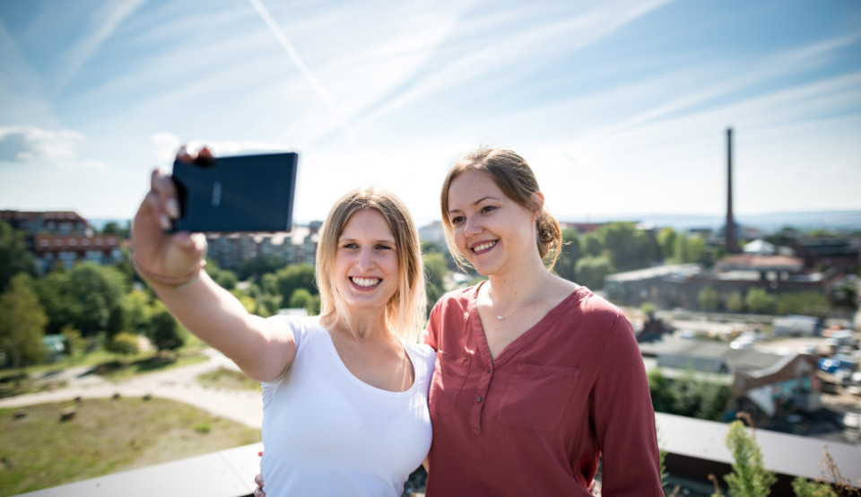 Zwei Hochschulmitarbeiterinnen machen ein Selfie auf einem Balkon Ihrer Hochschule.