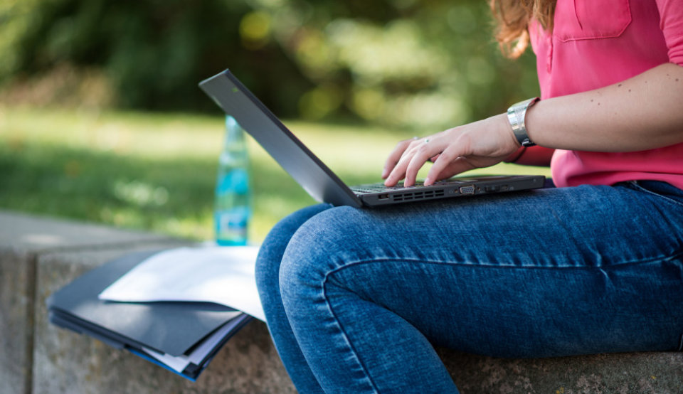Eine Frau sitzt draußen mit einem Laptop und schreibt auf der Tastatur