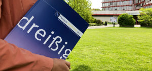 Eine Hochschulmitarbeiterin steht auf einer Wiese und hält den Erasmus+ Jahresbericht 2017 der NA DAAD in der Hand.